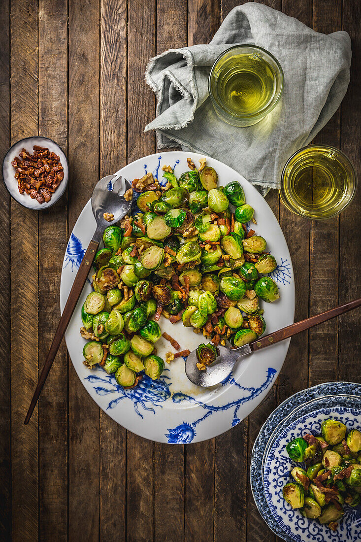 Steamed Brussels sprouts and bacon on a white and blue platter, with side plates and wine glasses