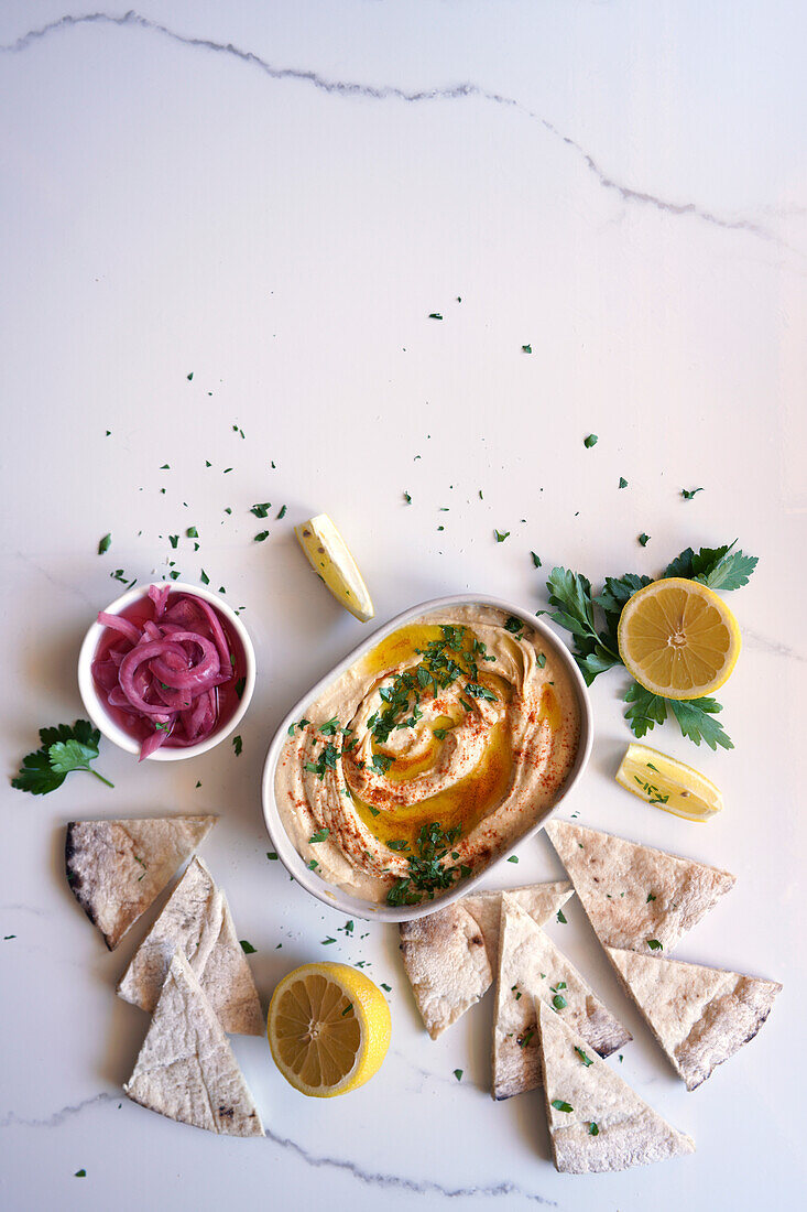 Chickpea hummus served with olive oil, pickled red onion, lemons, and flatbread on a white marble table background, with negative copy space.