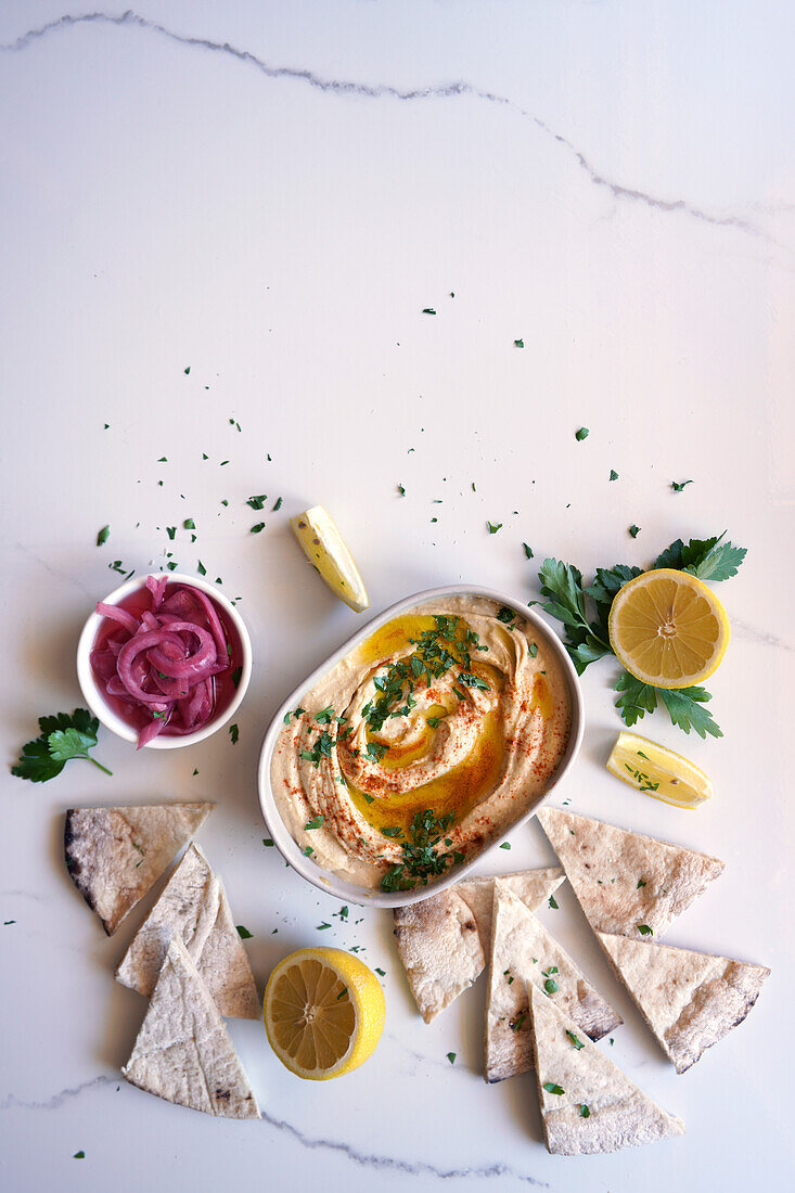 Chickpea hummus served with olive oil, pickled red onions, lemons and flatbread on a white marble table. Female hand dipping flatbread