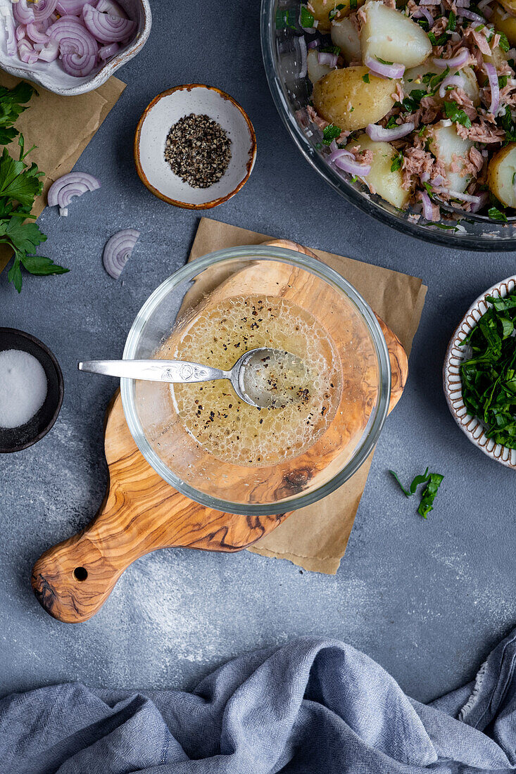 Salatdressing mit Zitronensaft und Olivenöl in einer kleinen Glasschale auf einem Holzbrett und Thunfisch-Kartoffelsalat an der Seite