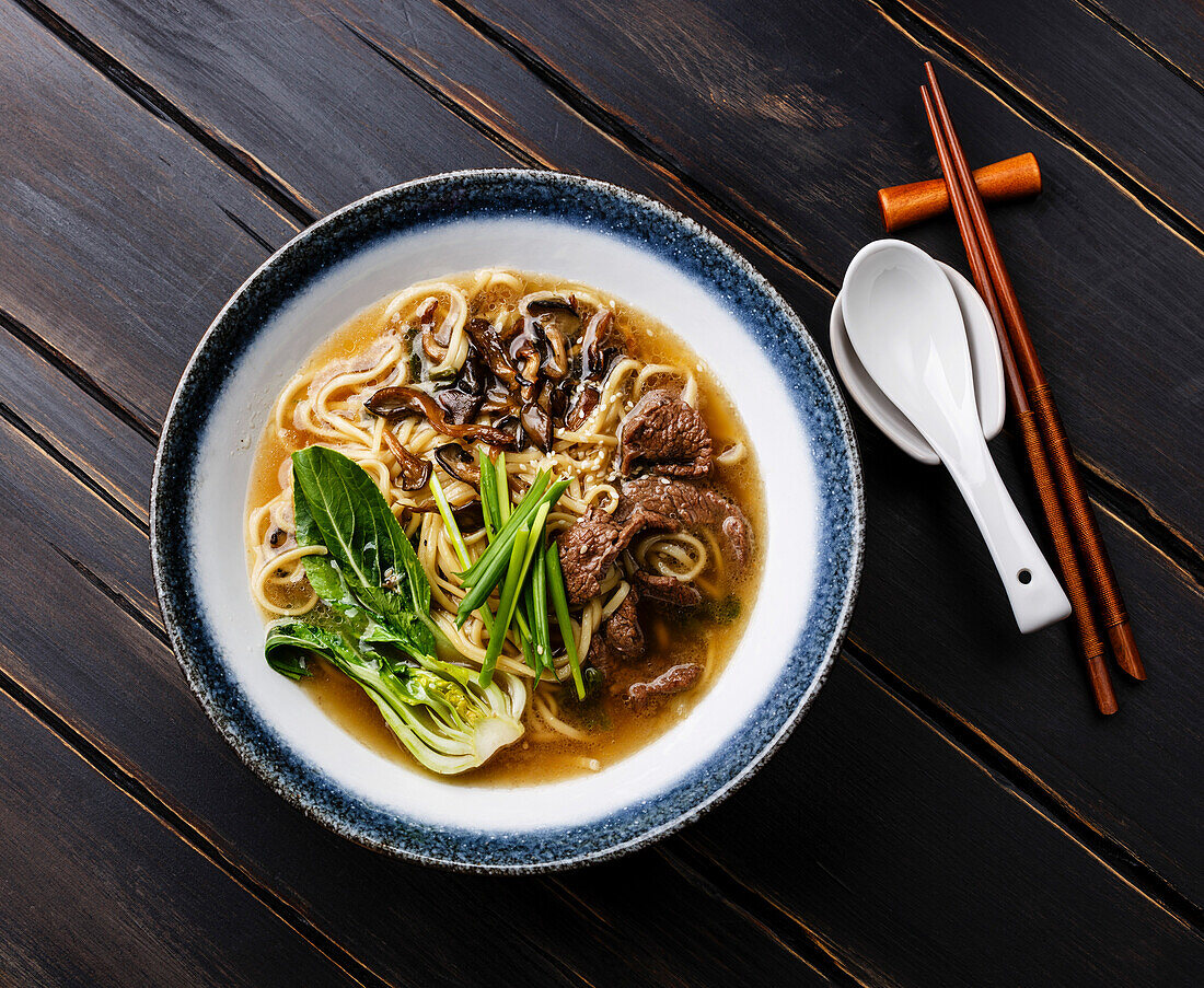 Ramen asiatische Nudeln in Brühe mit Rindfleisch und Pak Choi Kohl in Schüssel auf dunklem Holzhintergrund