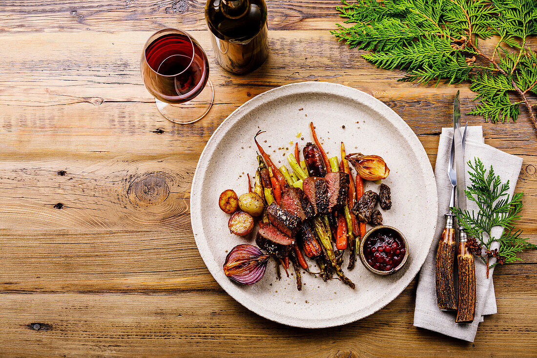 Gegrilltes Hirschsteak mit gebackenem Gemüse, Beerensauce und Rotwein auf Holzuntergrund