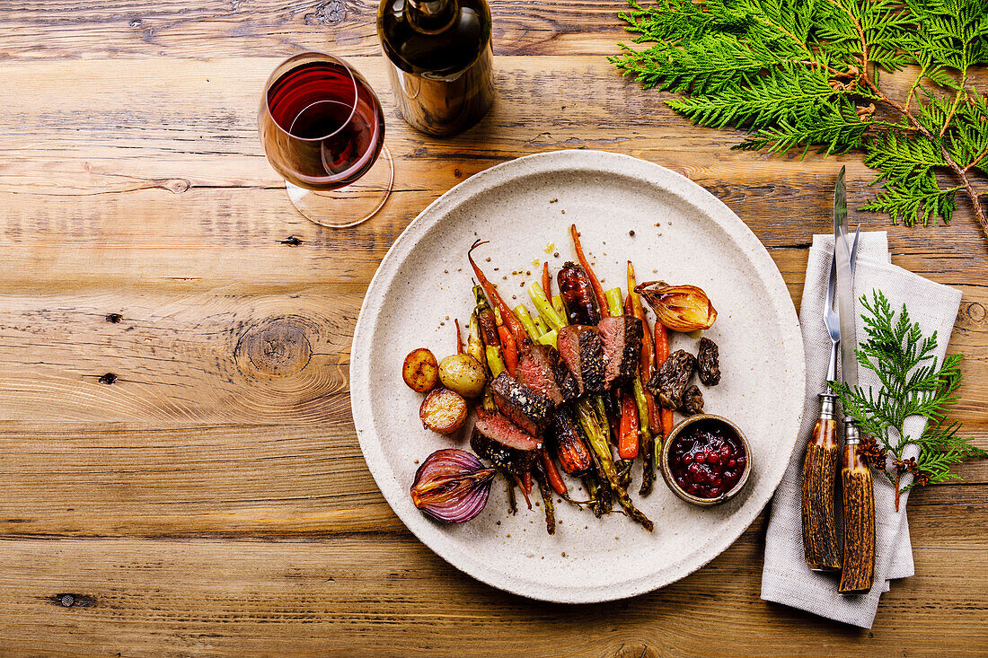 Grilled venison steak with baked vegetables, berry sauce and red wine on a wooden background