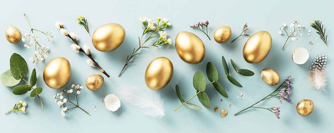 Easter composition of golden quail eggs, feathers and spring flowers on a pastel blue background. Spring holidays concept. Minimalist modern Easter background. Lay flat top view