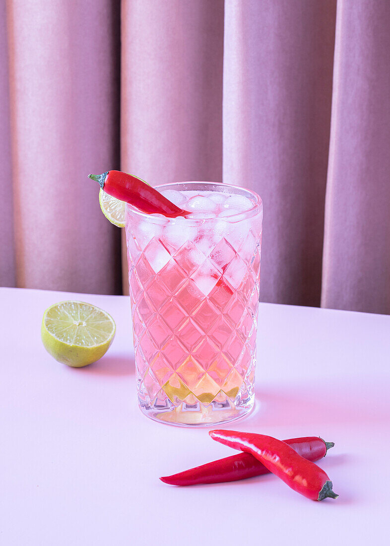 Clear glass filled with chilled refreshing cocktail, served with pepper and ice cubes on the table with lime and paprika