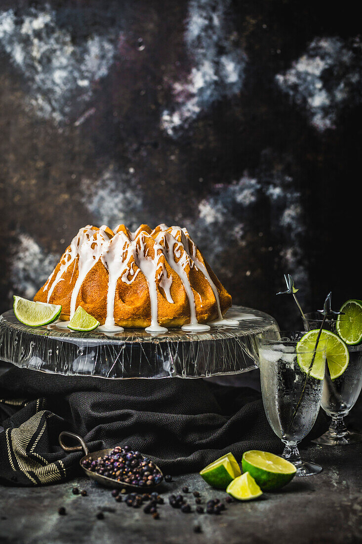 Glazed lime cake on a glass cake stand with spices, limes and cocktail glasses