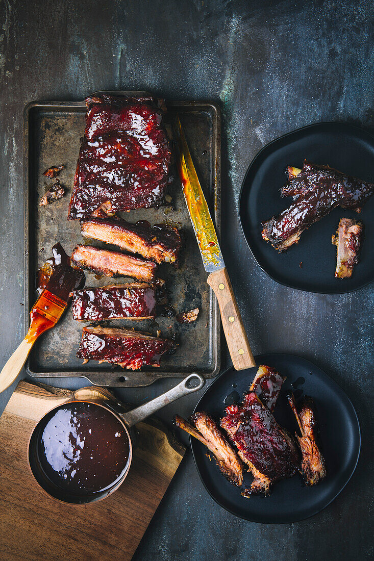 Glazed barbecued pork ribs, sliced and whole, on tray and two serving plates, with sauce pot and carving knife