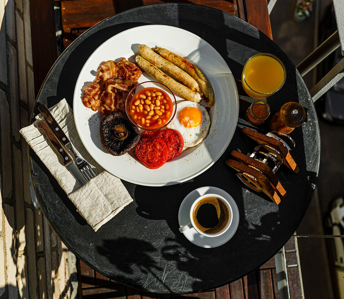 A full English breakfast with fried eggs, sausages, bacon, beans, toast and coffee, served on a round table on a sunny summer morning