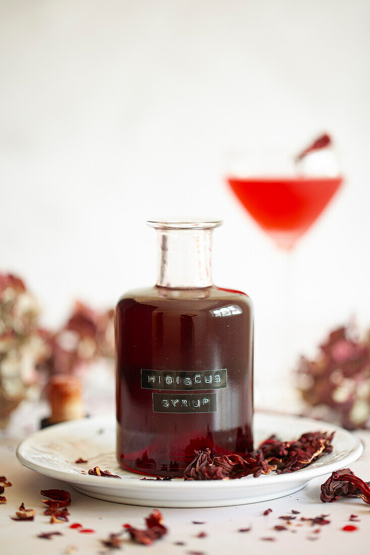 A bottle of homemade hibiscus syrup and a hibiscus cocktail in the background