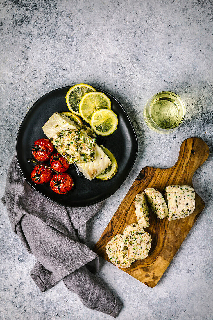 Top view of white fish fillet with melting butter on a black plate with fried tomatoes, sliced lemons and white wine in a stemless glass