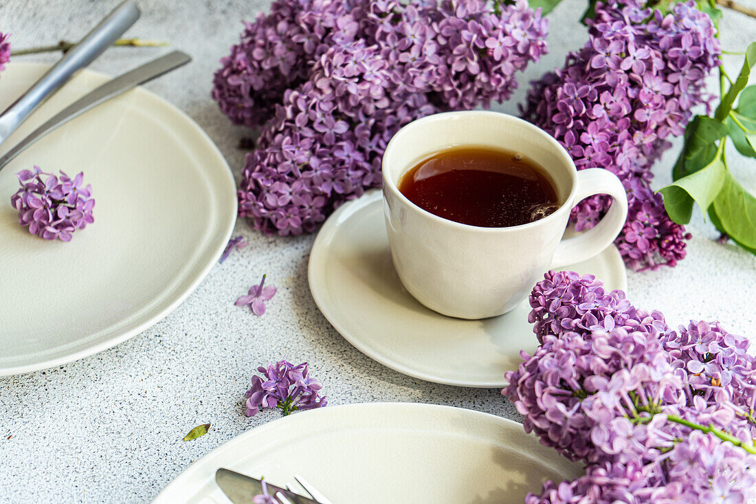Vorderansicht einer weißen Tasse Tee, weiße Teller, Messer und schöne fliederfarbene Blumen auf grauem Tisch