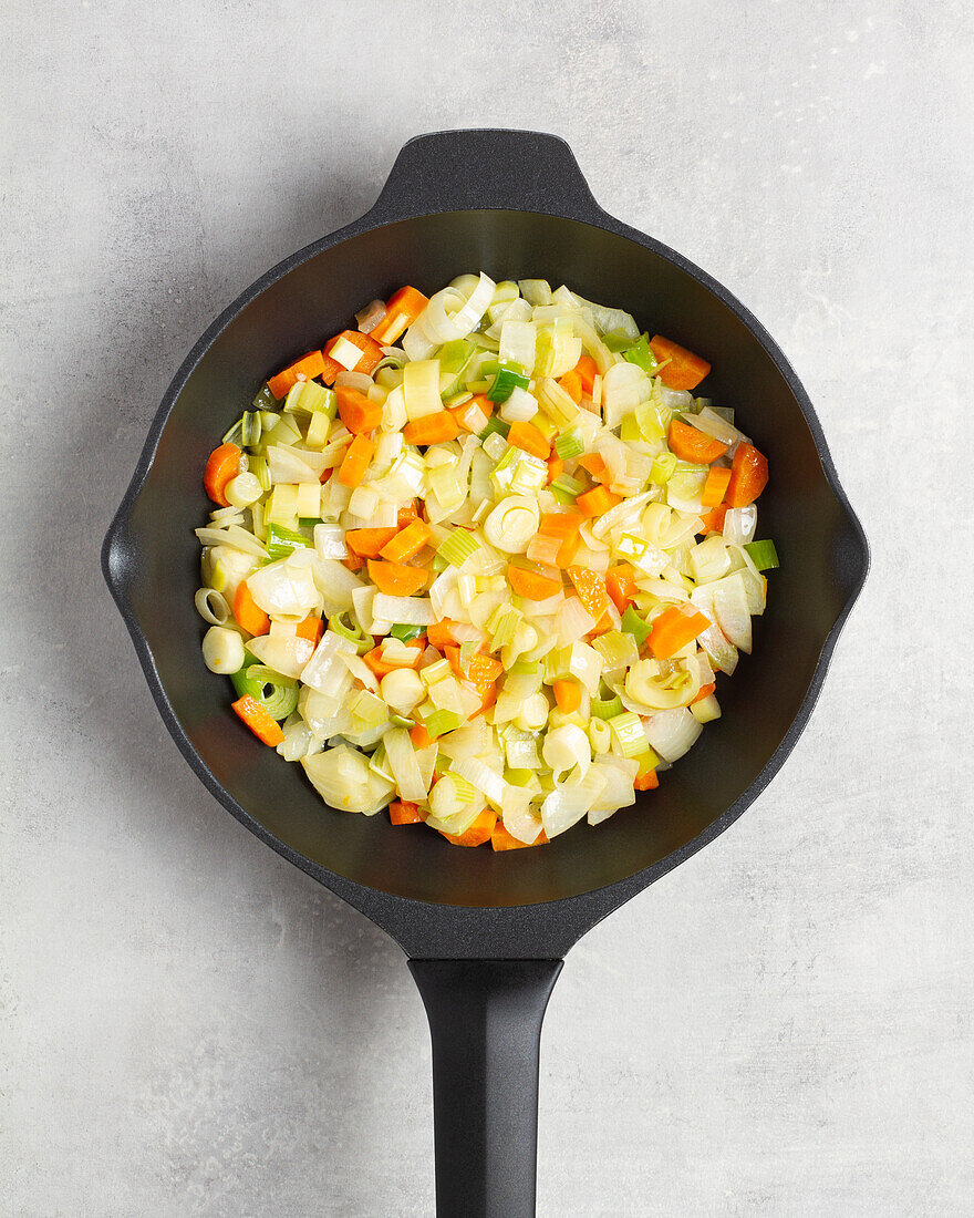 Top view of frying pan filled with fumet containing carrots cabbage and green strand placed on gray table in kitchen