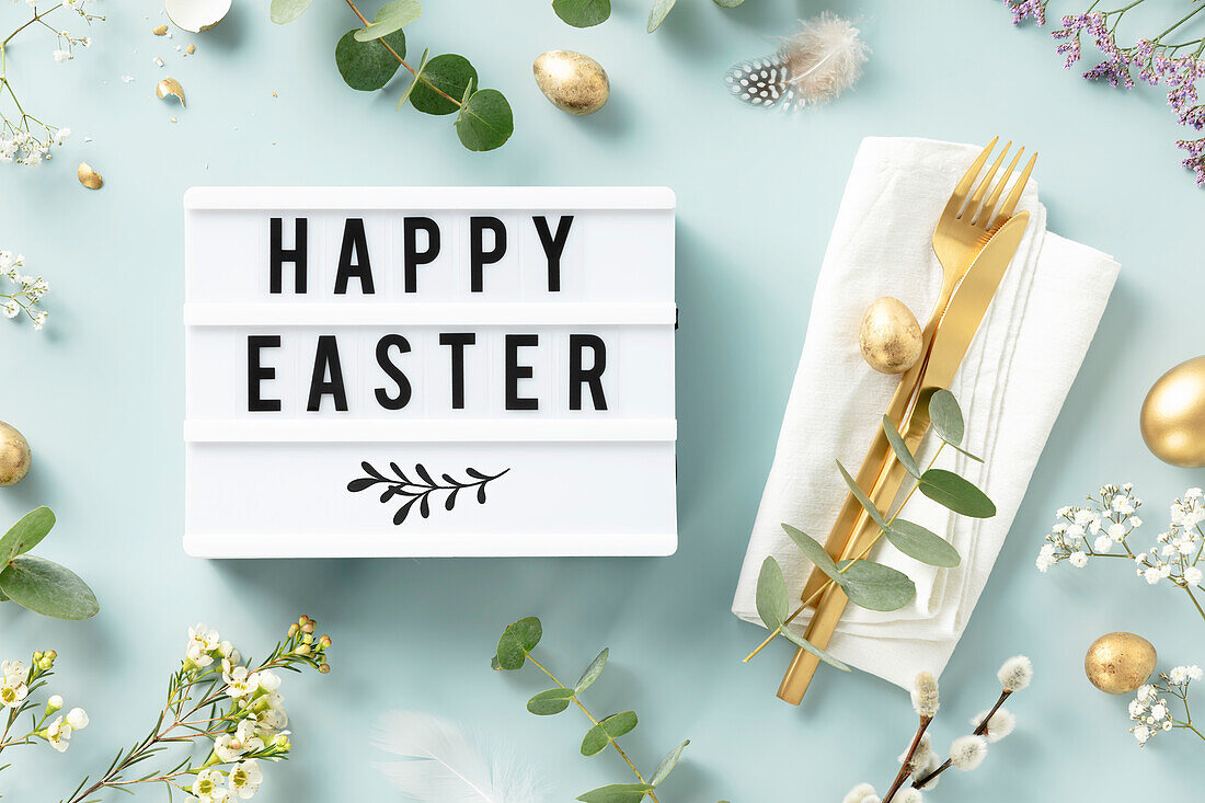 Easter table decorations. Stylish Easter brunch table setting with lightbox text Happy Easter, eggs, vintage cutlery, nests and spring branches on blue background top view flat lay
