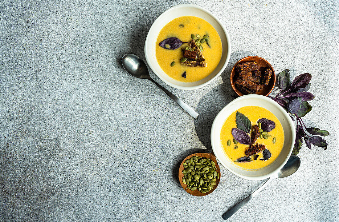 Top view of bowls of cream of pumpkin soup with basil herb, rye bread and seeds on blurred grey background with leaves