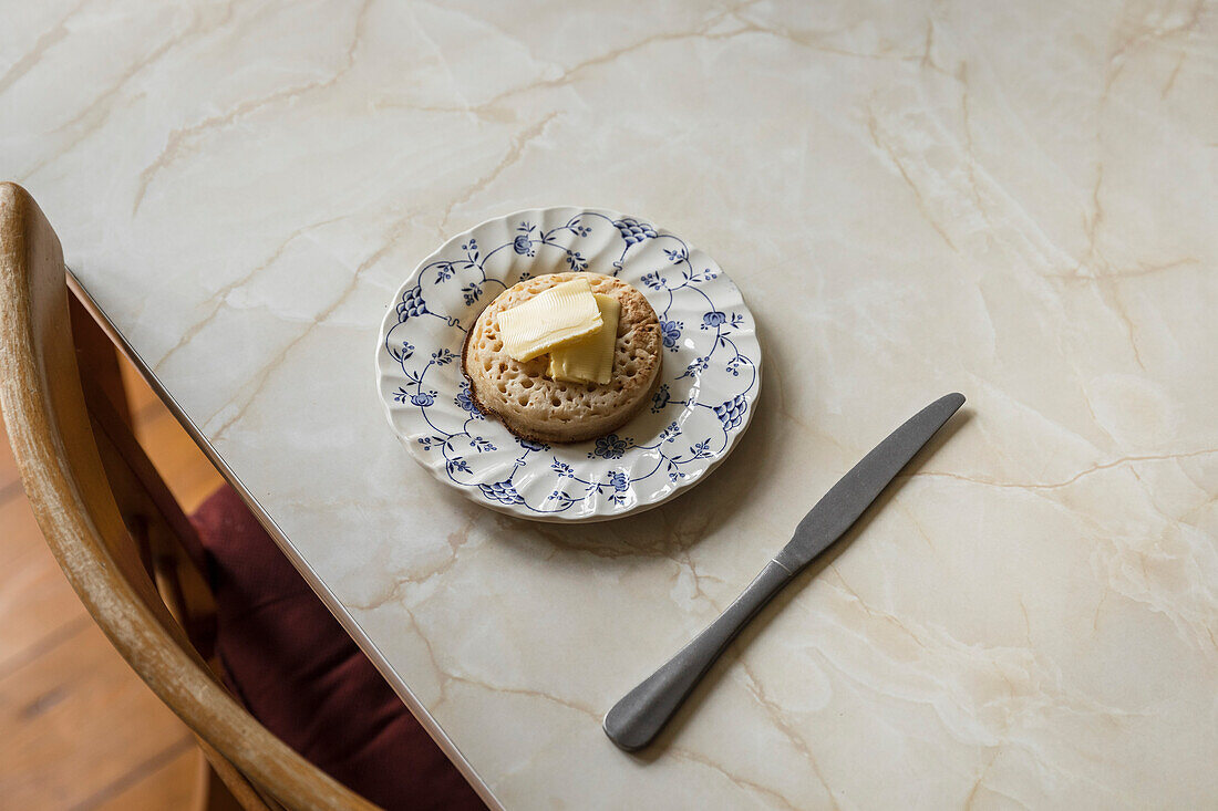 Crumpet with butter on blue floral plate on a marble table.