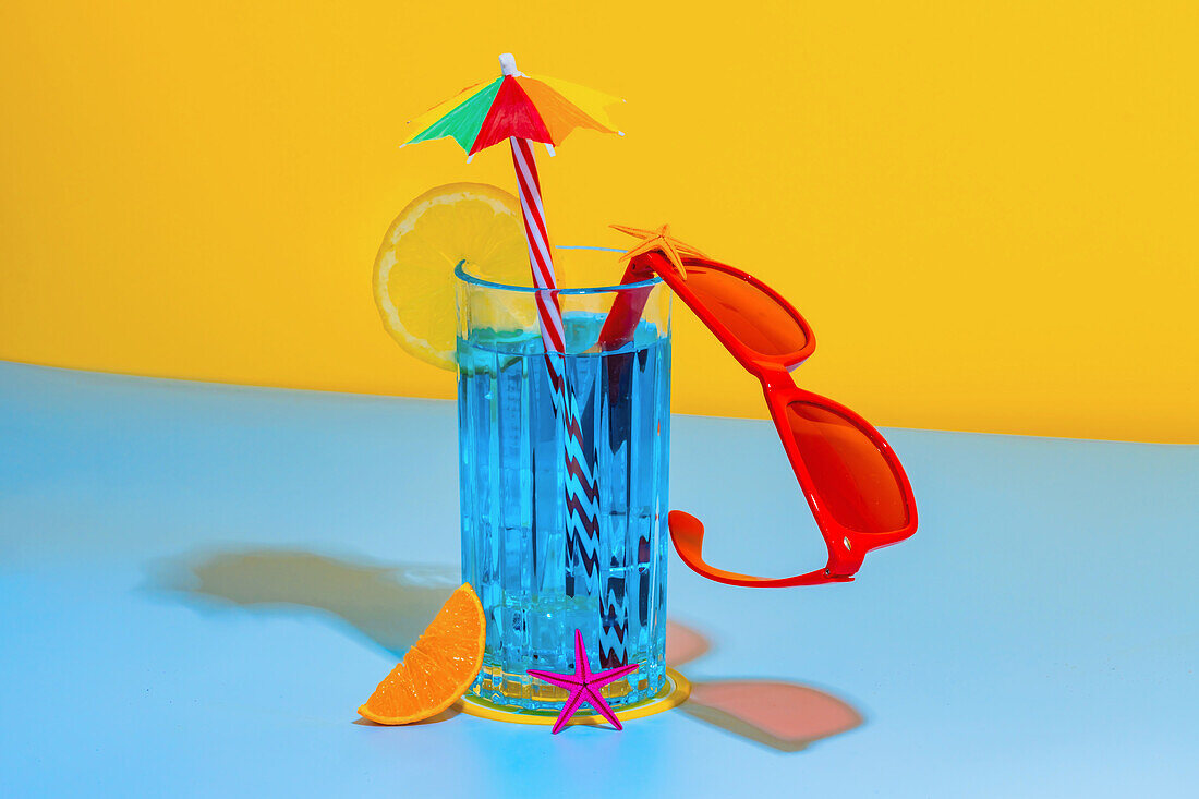 Composition of glass of blue liquid with slice of lemon and straw with stylish red eyeglasses placed on bright blue and yellow background in studio