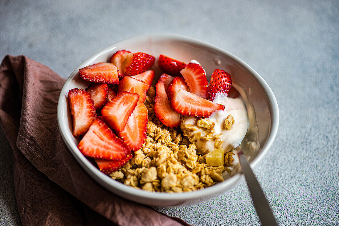 High angle of bowl with granola, yogurt and ripe organic strawberries with spoon on grey surface next to pink napkin against blurred background