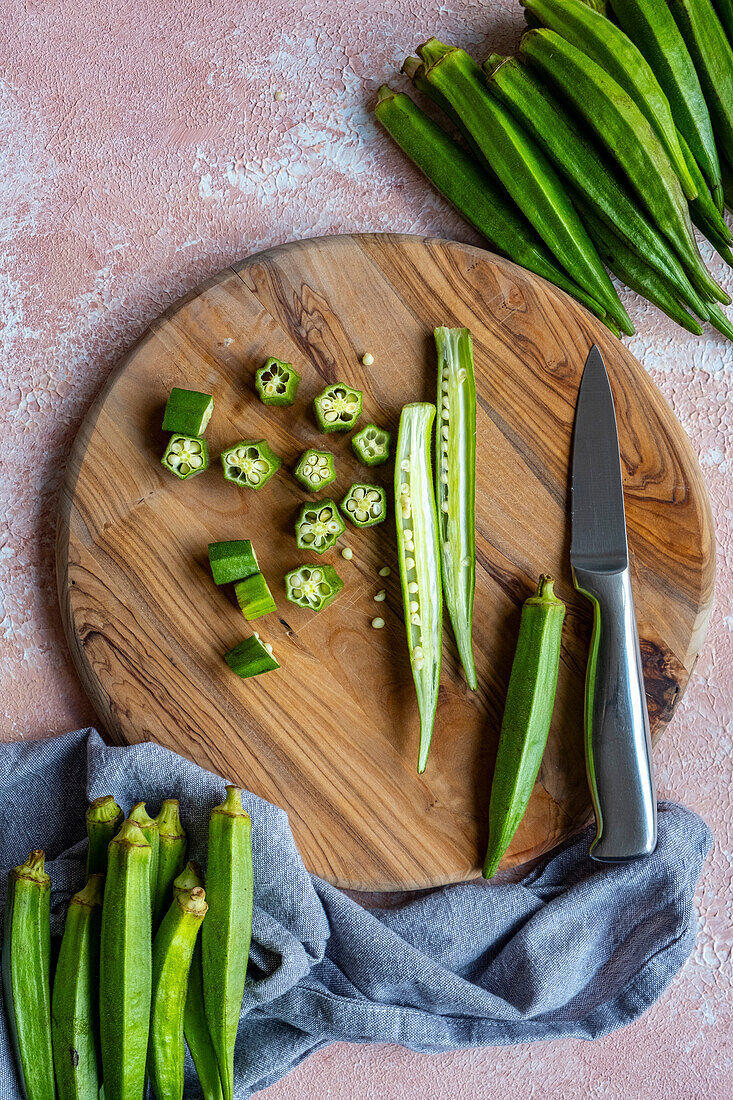 Fresh okra, sliced lengthways and crossways, on a wooden board and a knife on the side