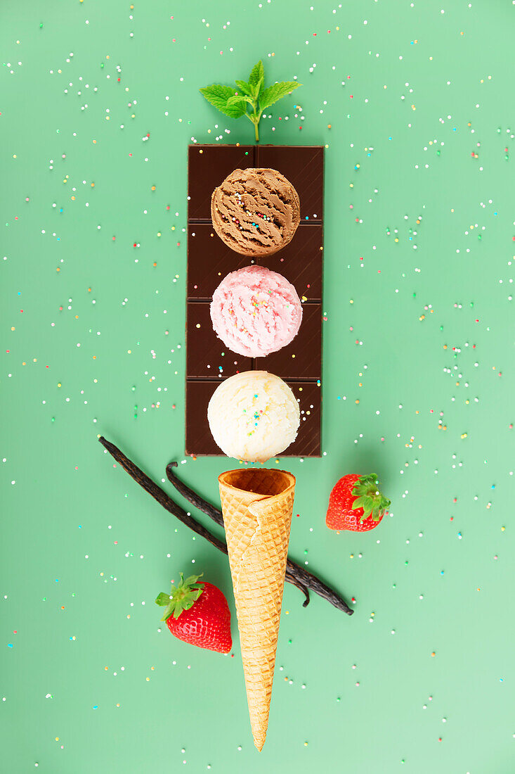 Chocolate, vanilla, strawberry scoops, waffle cone and ingredients on green background with colorful sprinkles, levitating concept. Spring or summer mood