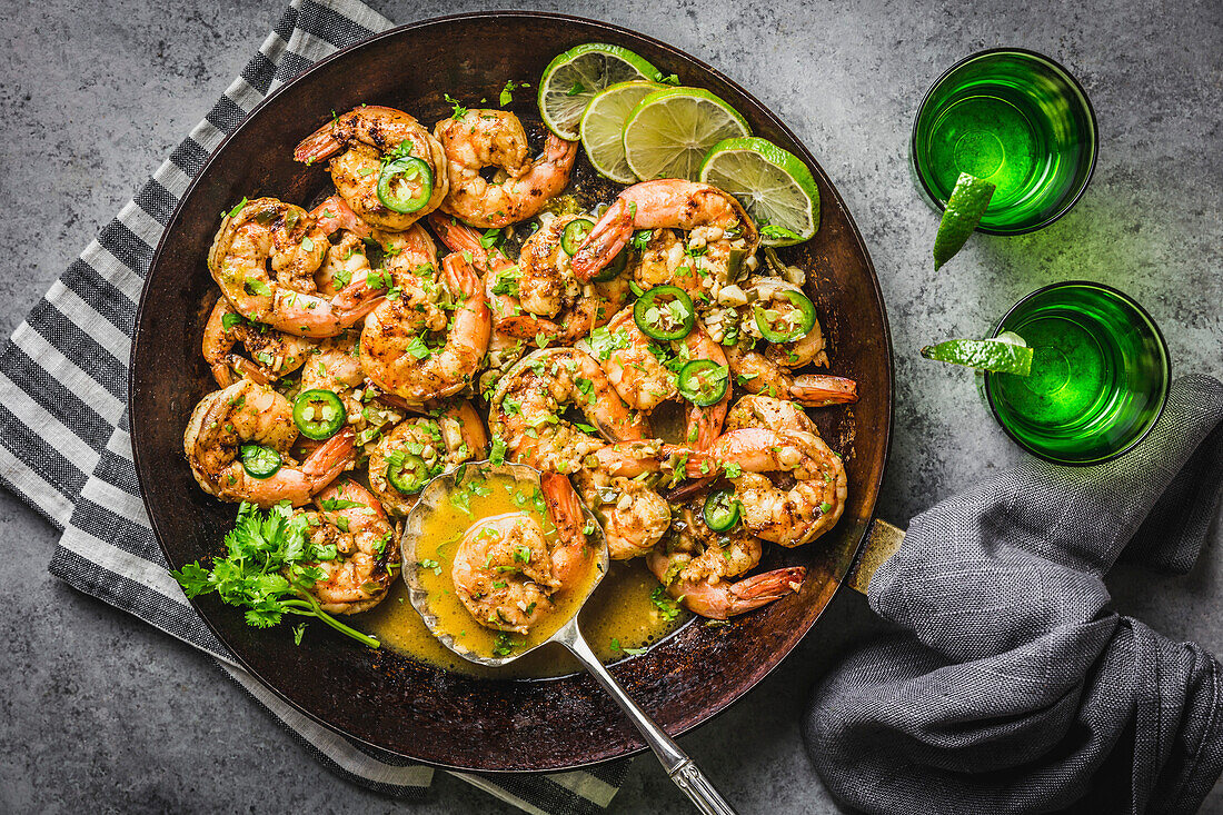 Shrimp with Chili, Lime and coriander in butter sauce, carbon steel skillet with gray linen napkins and silver spoon and green glass tequila glasses