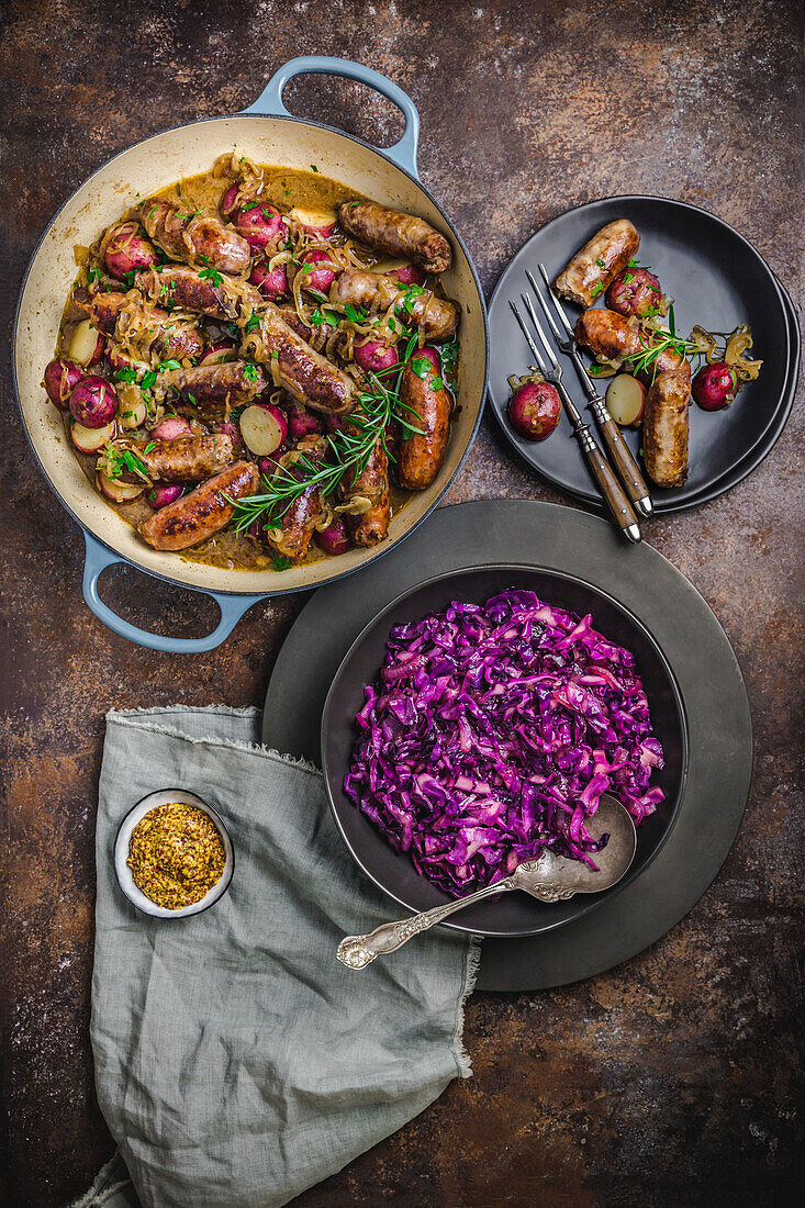 Niederländischer Ofen mit Würstchen, Kartoffeln, Kräutern, mit Schüssel mit leuchtendem Rotkohl