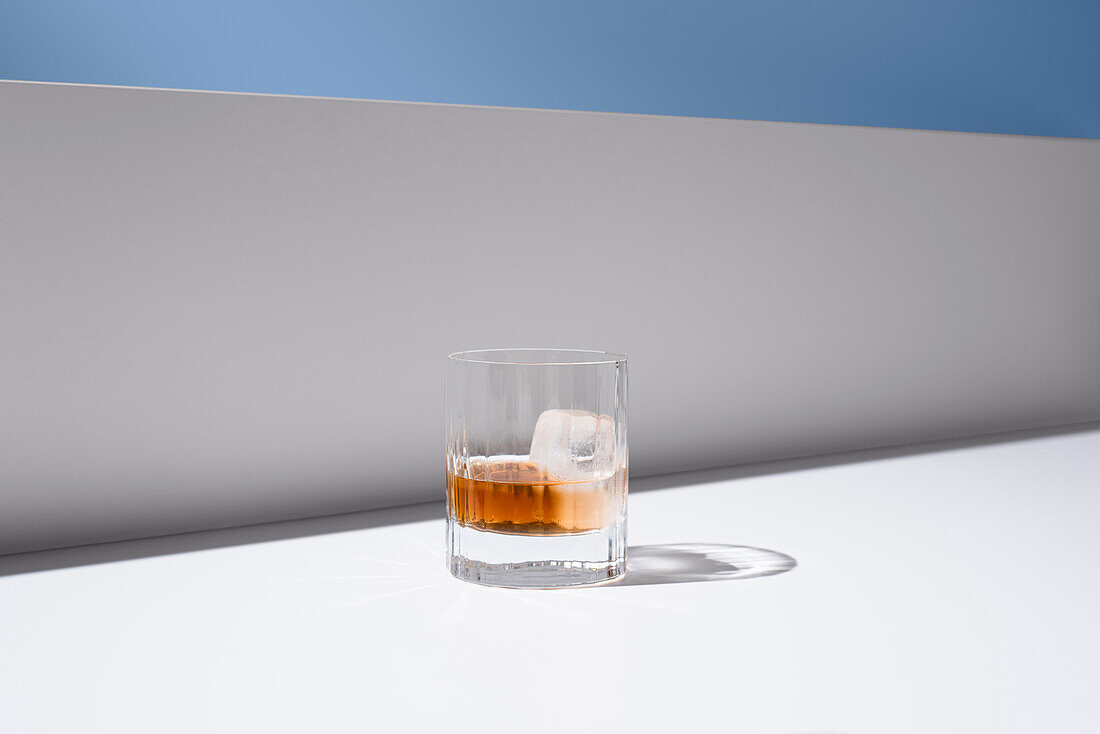 Kristallklares Glas gefüllt mit frischem altmodischem Cocktail, garniert mit Orangen und Eiswürfeln auf weißer Fläche zwischen weißen Wänden vor grauem Hintergrund