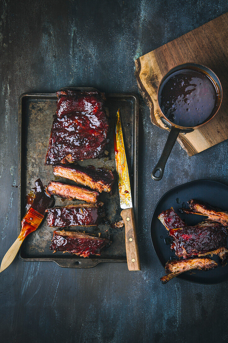 Glazed barbecued pork ribs, sliced and whole, on tray and one serving plate, with pot of sauce and meat knife