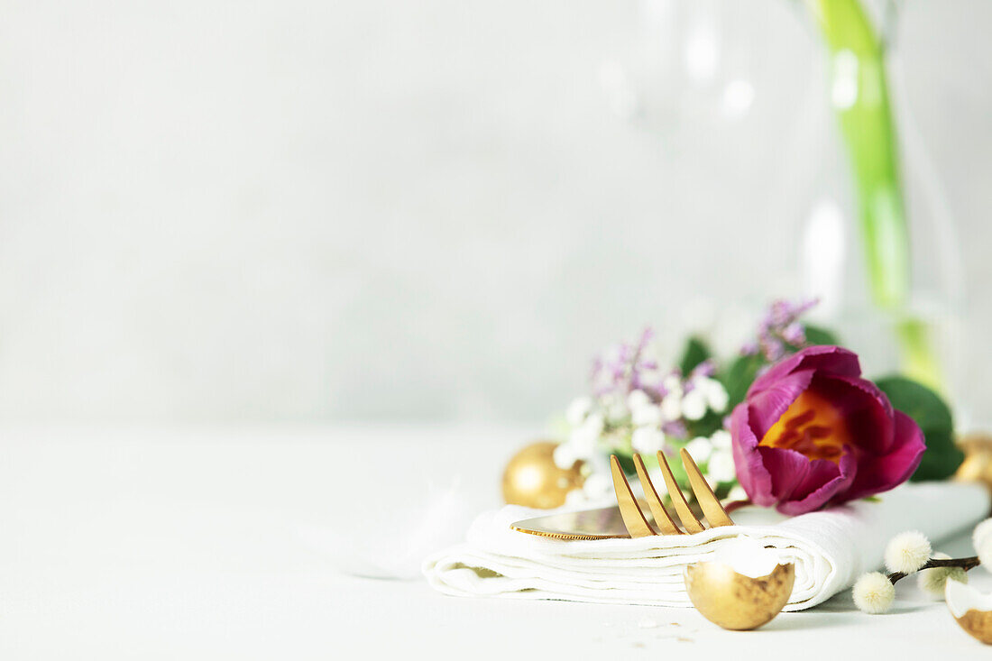 Easter table decoration with cutlery, spring flowers and golden eggs on a light grey background Copy space