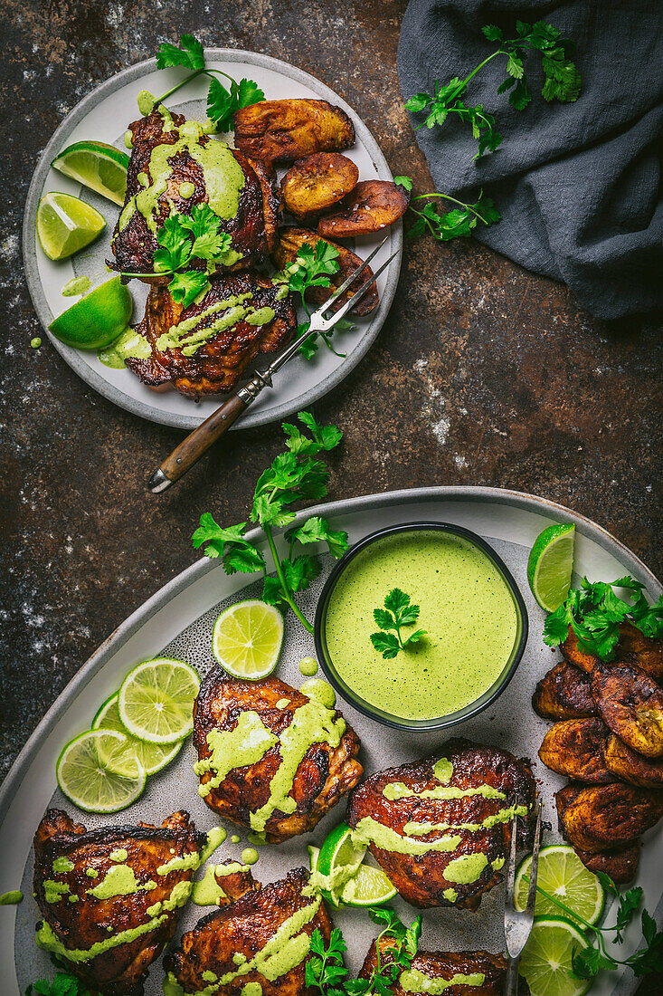 Peruvian chicken thighs on a platter and serving dish, with fried plantains, green sauce and lime and herb garnish