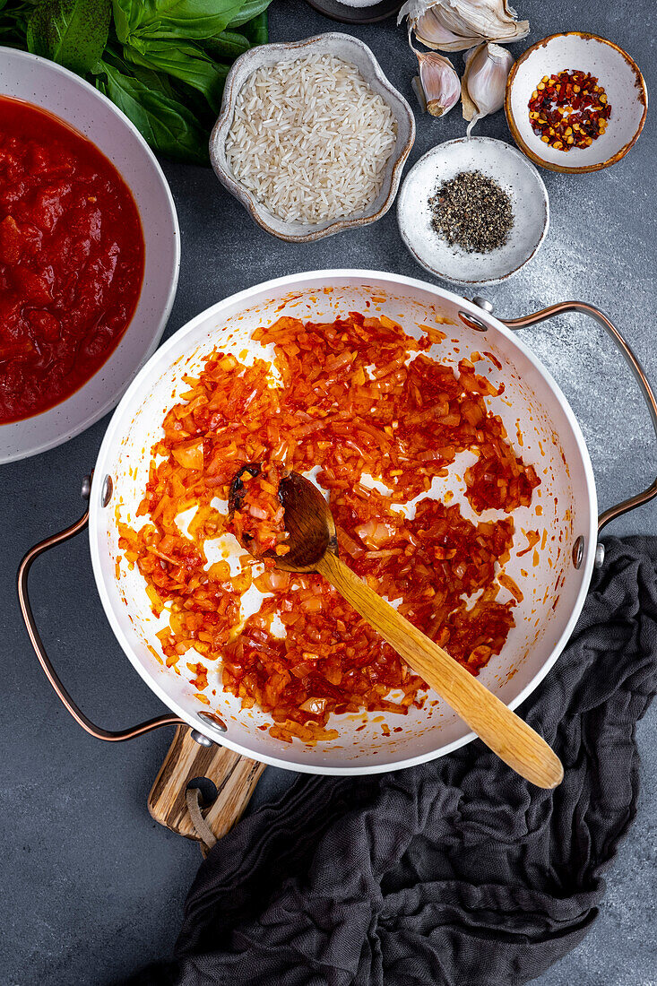 Diced onion beind cooked with tomato paste in a white pot, a wooden spoon inside it, chopped tomatoes, spices, rice, basil and garlic cloves on the side.