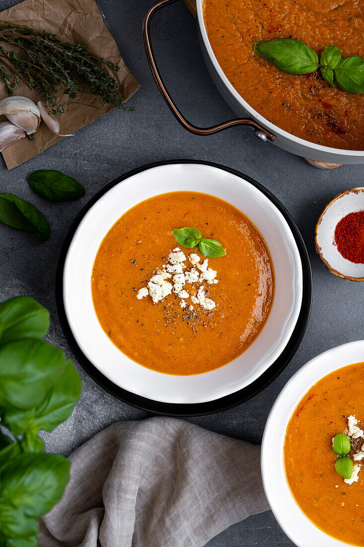 Roasted tomato soup garnished with feta cheese and basil in two white bowls and the soup pot on the side