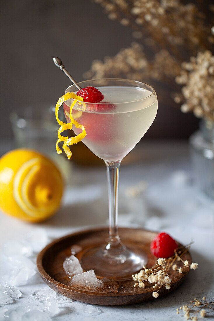 A vodka and limoncello cocktail in a Nick and Nora glass, garnished with lemon zest and fresh raspberries