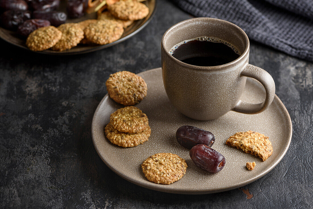 A cup of coffee on a plate with dates and cereal cookies.