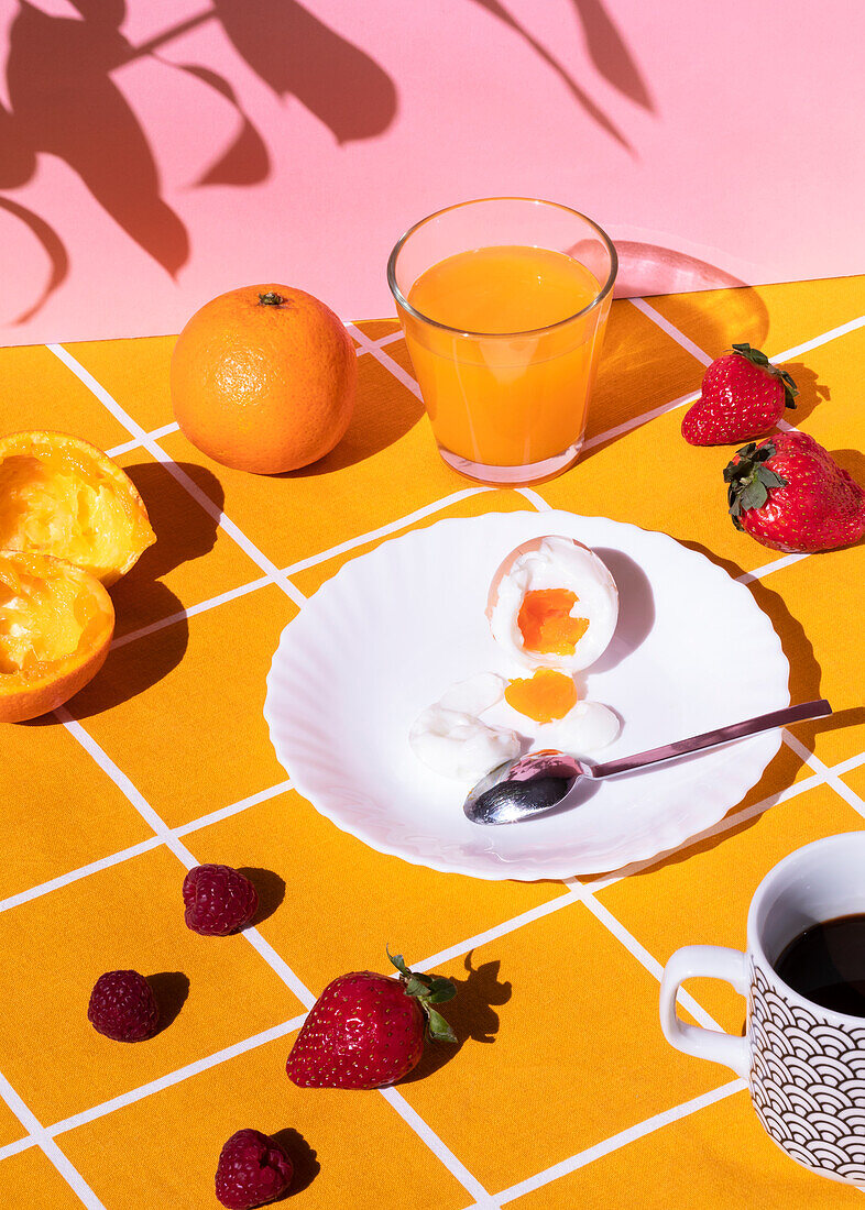 Hoher Blickwinkel auf ein leckeres Frühstück mit gekochtem Ei, umgeben von Erdbeeren und Himbeeren mit Orangensaft und einer Tasse Heißgetränk am Morgen