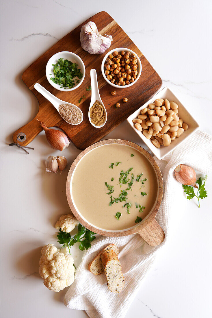 Vegane Suppe aus Blumenkohl, Kreuzkümmel und Limabohnen mit Zutaten auf weißem Marmorboden. Flatlay von oben nach unten
