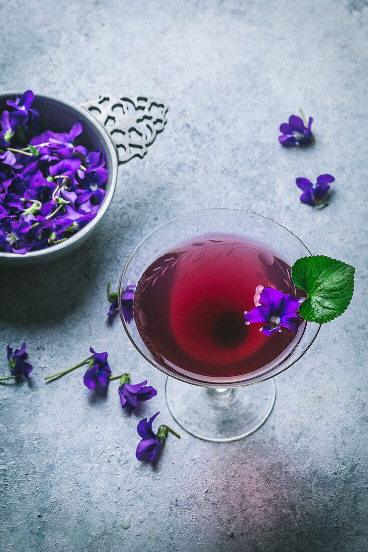 Purple cocktail in a vintage glass with flower garnish and petals in a bowl on a grey concrete background