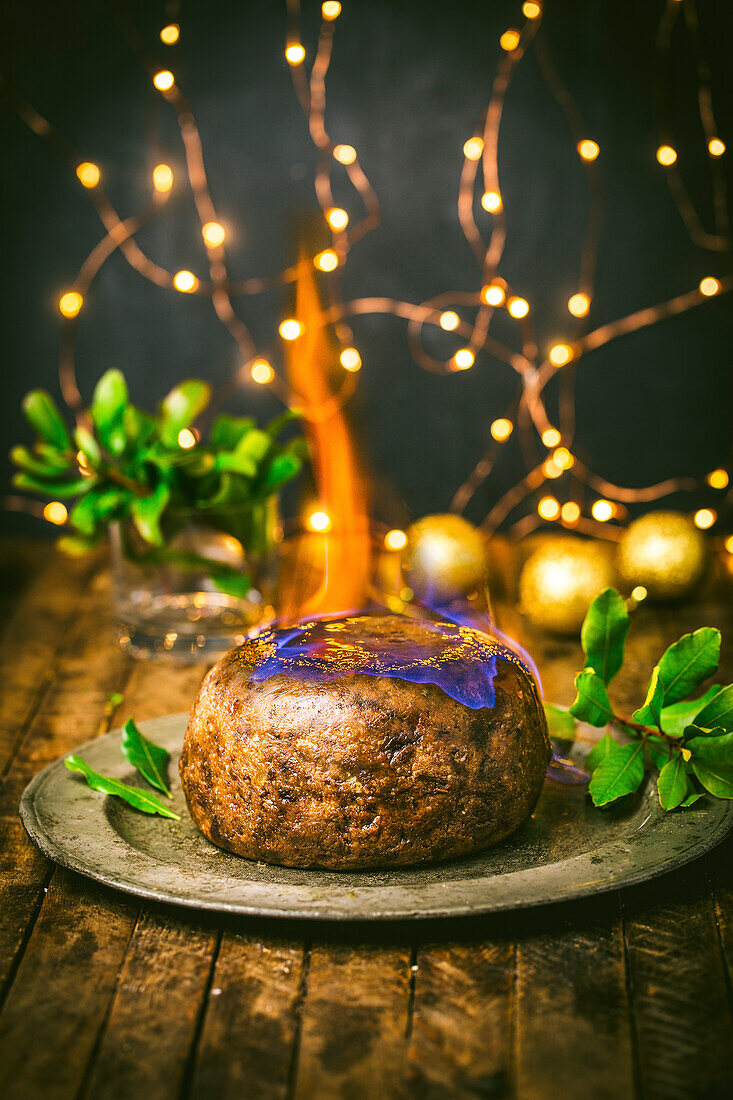 Christmas pudding with flame on pewter plate with holly and Christmas lights