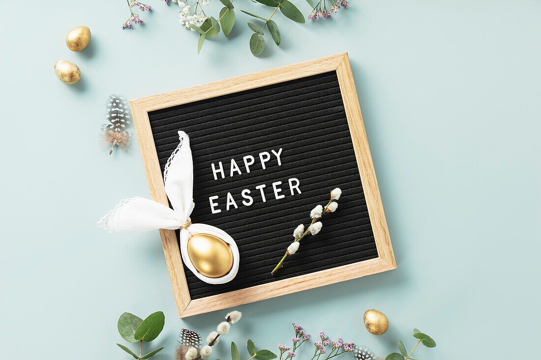 Stylish Easter flat lay with letter board, egg in easter bunny napkin, golden eggs, feathers and spring flowers on blue background. Minimalist modern Easter background. Top view flat lay