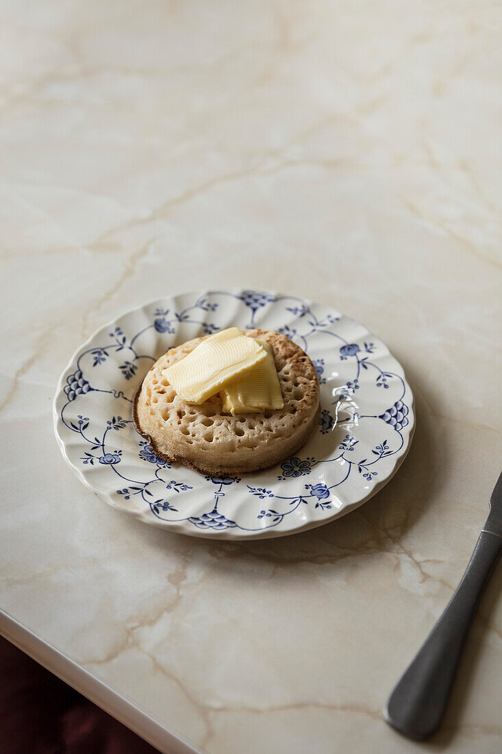 Crumpet with butter on a blue flower plate on a marble table