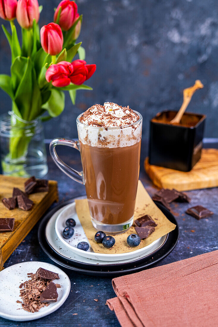 Heiße Schokolade mit Schlagsahne in einem Latte-Glas, Blaubeeren und Schokoladenstückchen drum herum, dahinter Blumen