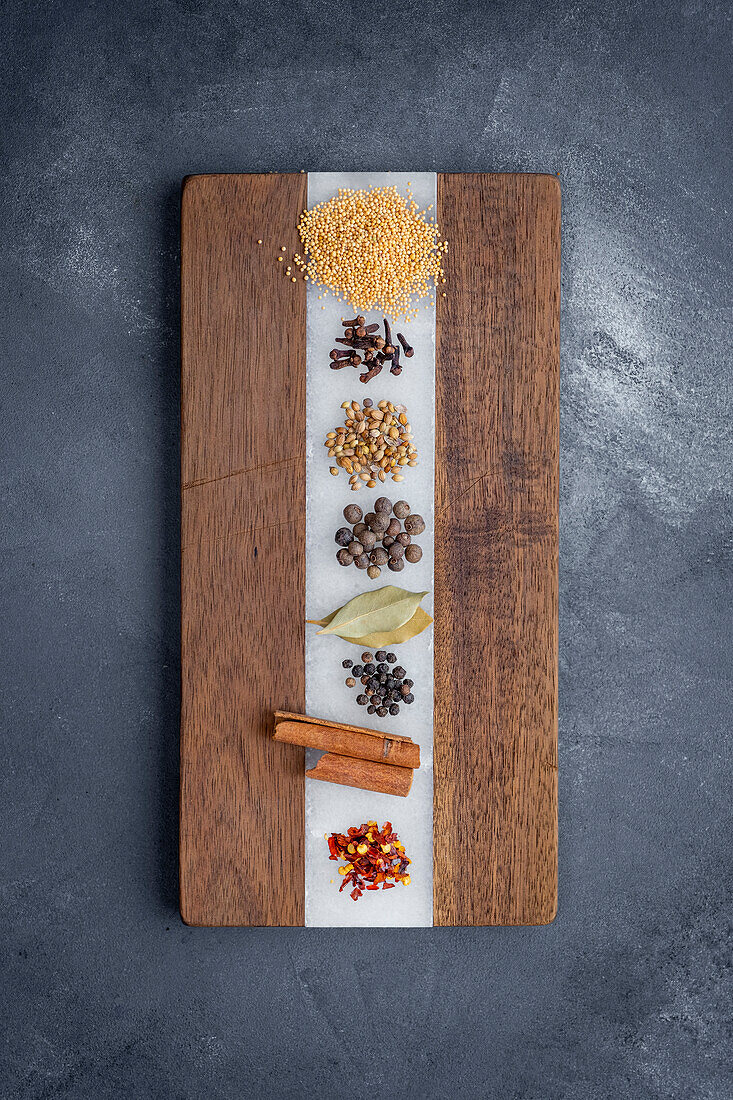 Coriander seeds, cloves, mustard seeds, black peppercorns, bay leaves, cloves, cinnamon sticks, red pepper flakes photographed on a wooden chopping board