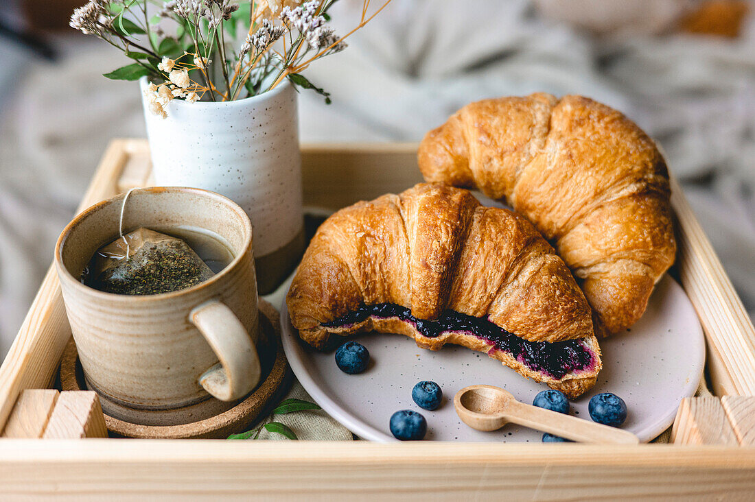 Frühstück im Bett mit frisch gebackenen Croissants mit Blaubeermarmelade und Tee in einem Becher