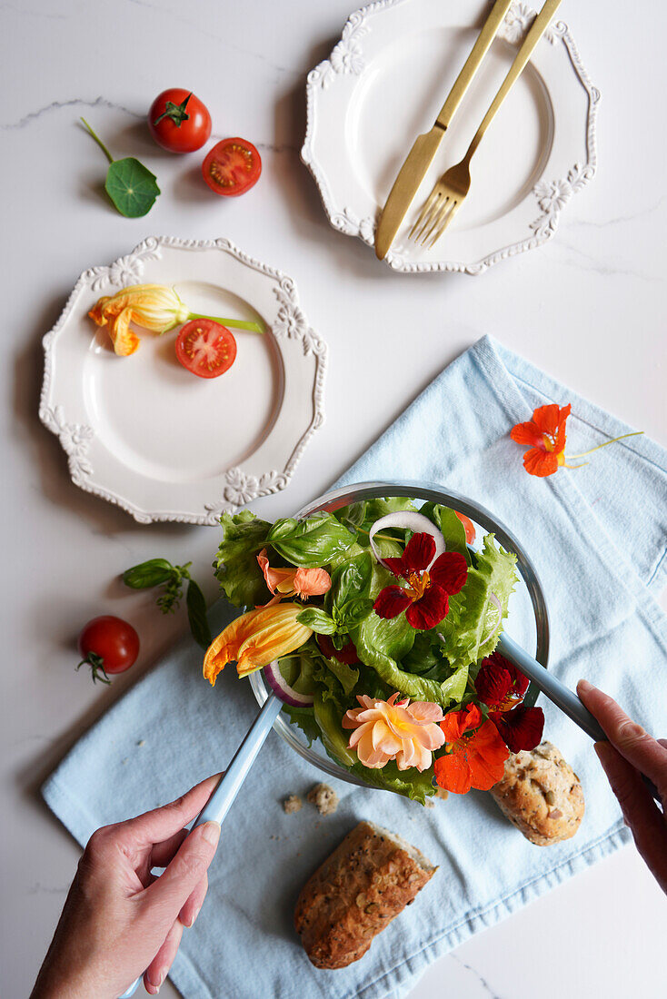 Woman's hands serving Mediterranean salad with zucchini, nasturtium, and rose edible flowers flatlay.