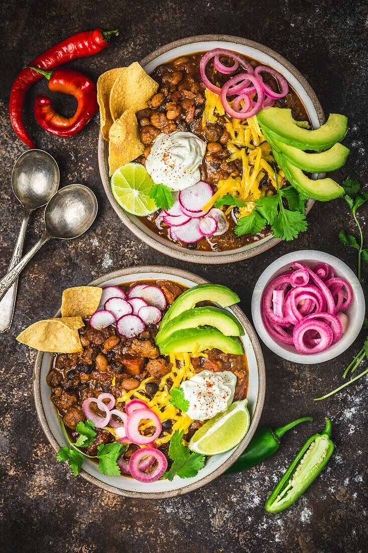 Chilli con carne in two bowls with garnishes, pickled red onions and spoons in bubbles on a dark background