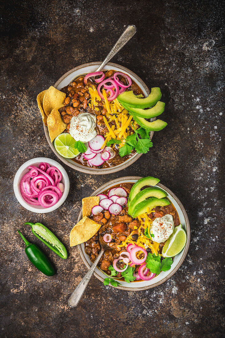 Chili con carne in zwei Schalen mit Beilagen, eingelegten roten Zwiebeln und Löffeln in Blasen auf dunklem Hintergrund