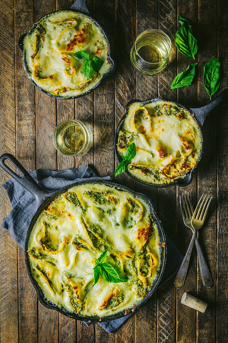 Two small and one large cast-iron pan with baked cheese spaetzle, wine and basil garnish on wooden table