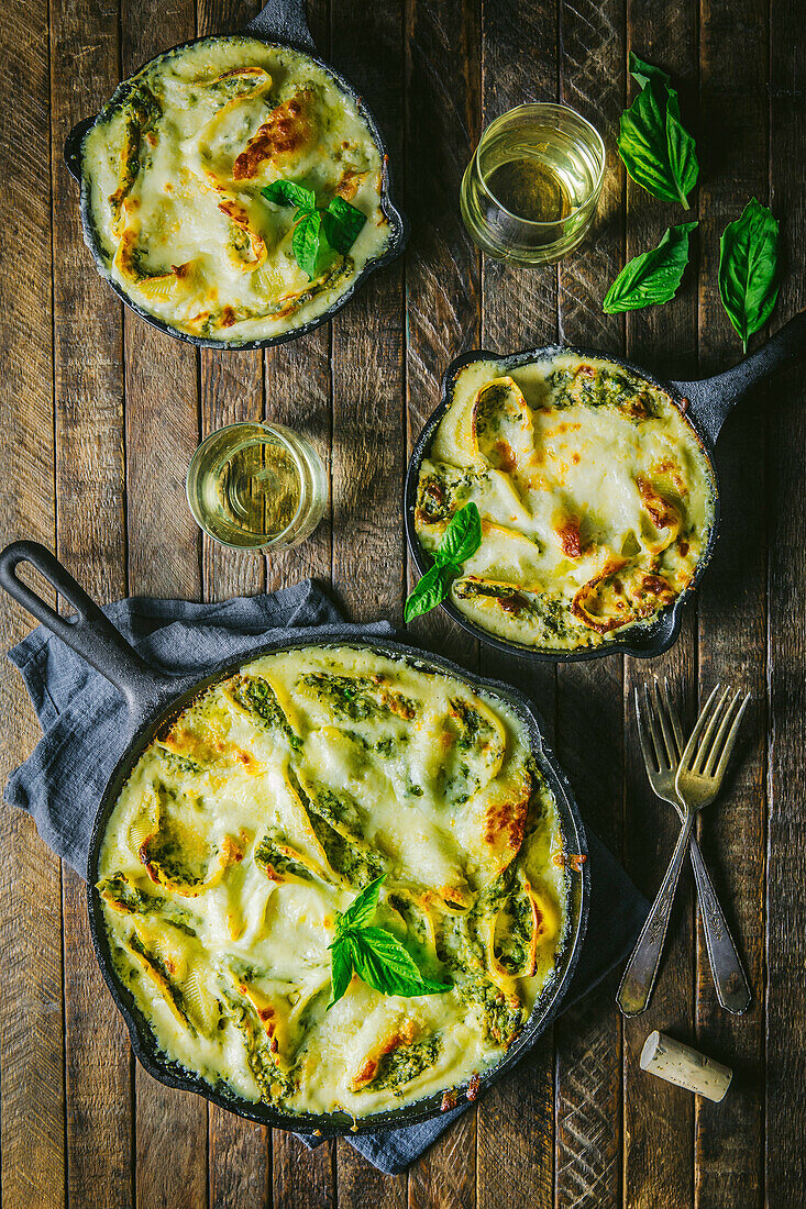 Zwei kleine und eine große gusseiserne Pfanne mit gebackenen Käsespätzle, Wein und Basilikumgarnitur auf Holztisch