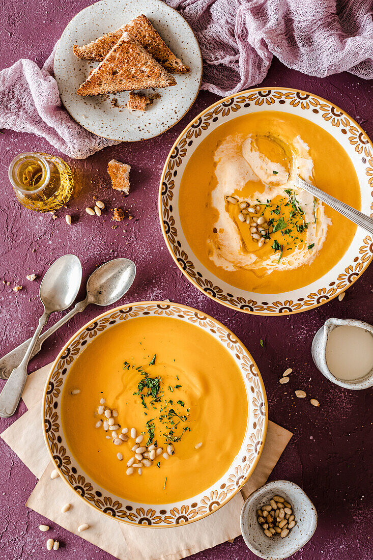 Two bowls of pumpkin soup served with brown toast and garnished with cream, pine nuts and herbs on a purple background