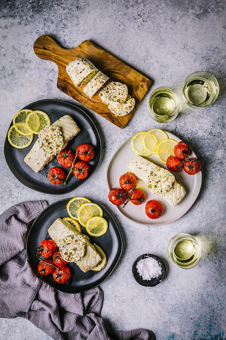 Overhead wide shot with White fish filets topped with firm compound butter on 2 black, one white plates with roasted tomatoes, sliced lemons and white wine in stemless glasses