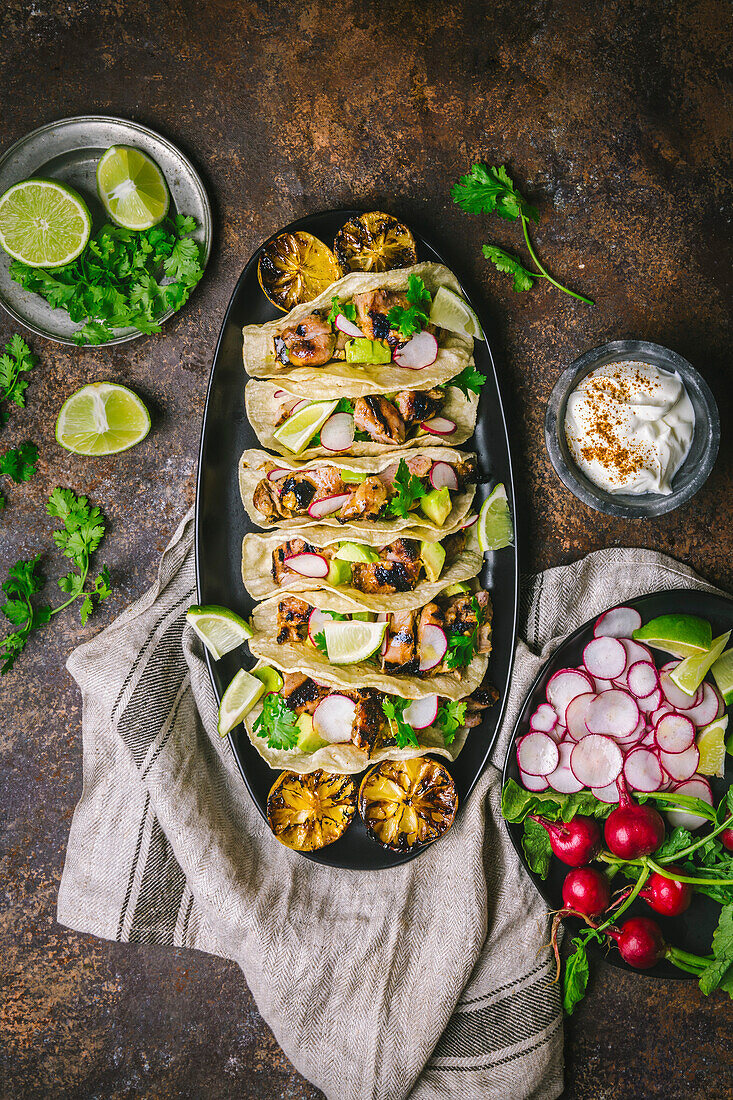 Arrangement of Grilled Chicken Tacos on a dark oval platter with sliced radishes, limes, coriander, charred lemons and sour cream